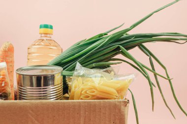 Muhtaçlar için bağış kutusu. Tereyağı, konserve yiyecek, soğan, kurabiye, makarna ve meyveli pembe bir zemin üzerine izole edilmiş karton kutu. Yemek teslimatı.