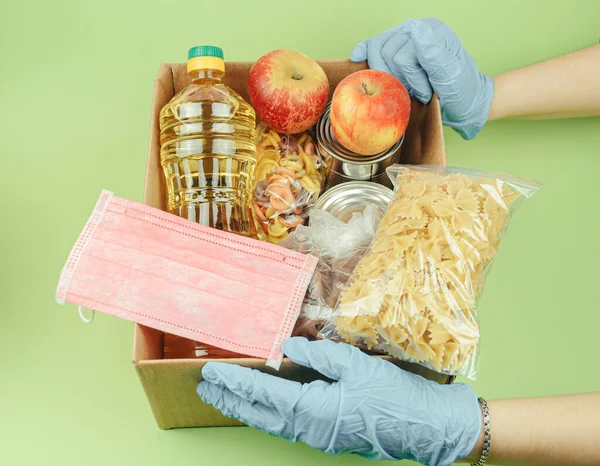 Volunteer in protective gloves with a donation of food boxes on a green background. Food delivery and virus protection. Donation and mercy concept.