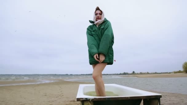Het meisje in een oude jas staande in een badkuip op de rivieroever. — Stockvideo