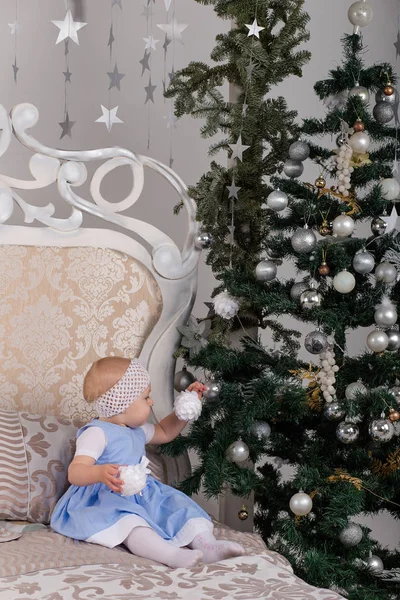 The little girl in a blue dress on the bed at the Christmas tree — Stock Photo, Image