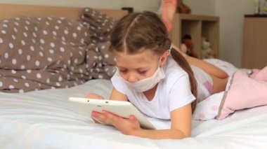 Solunum maskeli küçük bir kız tablette karantinada oturuyor.