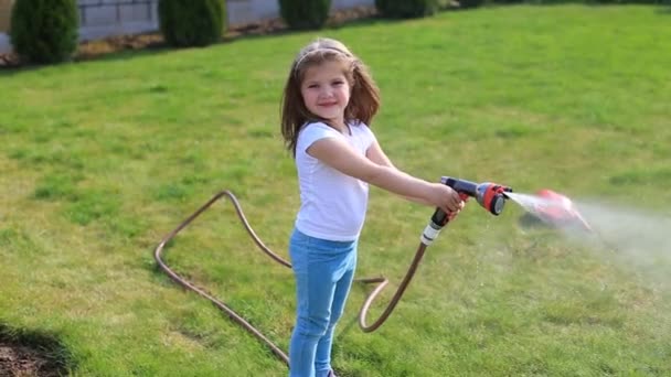 Liten flicka med en slang i händerna vattna en grön gräsmatta — Stockvideo