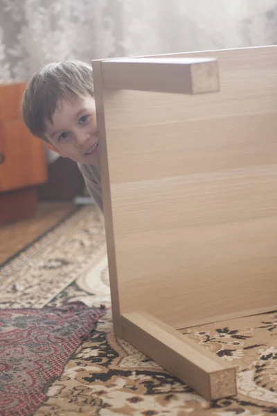 Divertido chico se esconde detrás de los muebles de interior en la habitación . —  Fotos de Stock