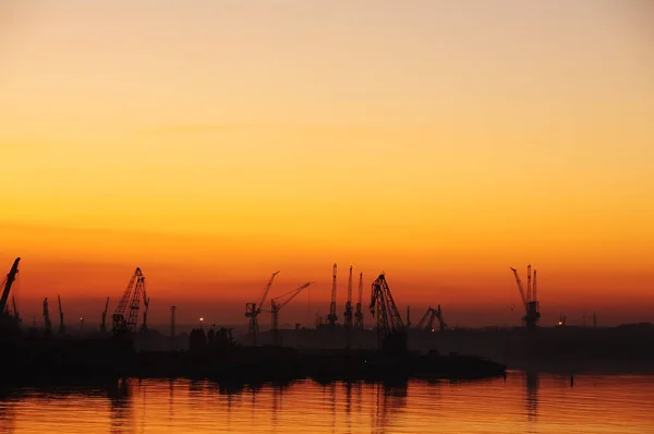 Silhouettes of cranes in the port and reflection in the water at sunset. Dark photo.