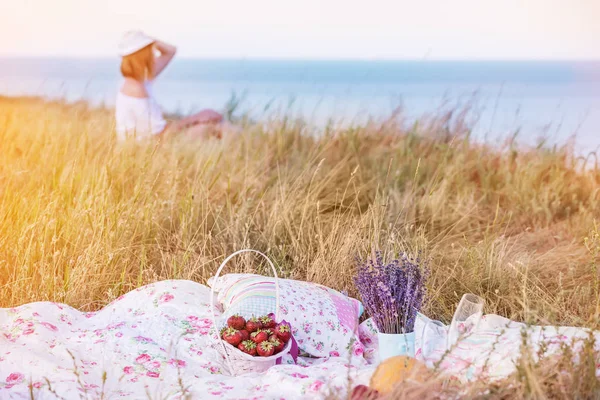 Picnic on a cliff near the sea, fruits on a colorful blanket and faded figras of a girl sitting in the grass on a cliff overlooking the sea.