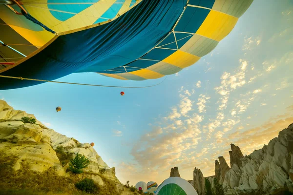 A huge balloon is inflated by fire, preparing for flight. in the background are other balloons and beautiful rocks.