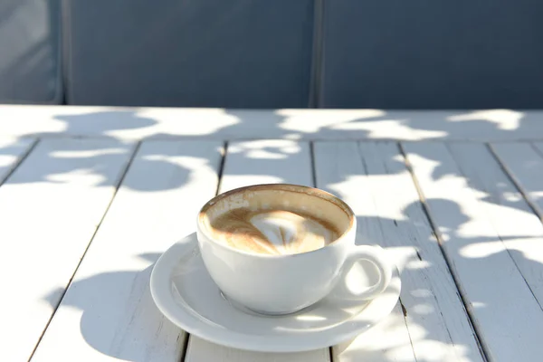 Cup with a cappuccino on a white wooden table on the street. A play of shadows on a wooden surface.