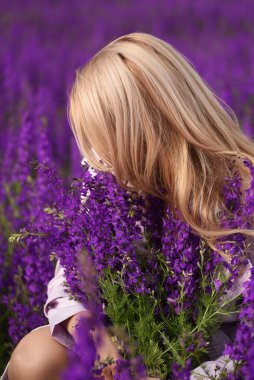 Sarışın, uzun saçlı, çiçek açan leylak çiçeklerinin arasında elinde buket olan bir kadın. Yüzü olmayan düşünceli bir fotoğraf..