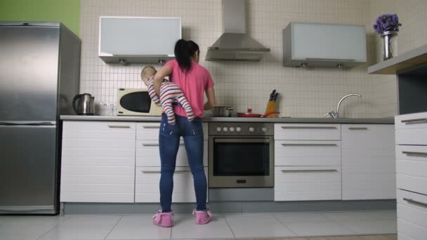 Ocupada madre cocina en la cocina sosteniendo bebé hijo — Vídeo de stock