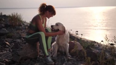 Gün batımında kıyıda oturan zinde bir kadın köpeği okşuyor.