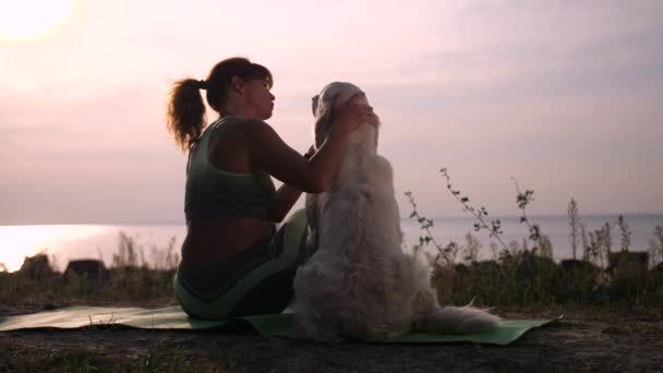 Gün batımında deniz kıyısında köpeğini okşayan dişi sporcu. — Stok video