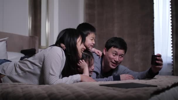 Улыбающаяся китайская семья делает селфи лежа на кровати — стоковое видео
