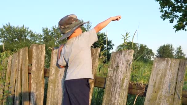 Cute boy in funny hat standing on village fence — Stock Video
