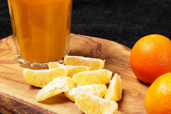 Vários cítricos maduros e um copo de suco em uma mesa de madeira em um quadro-negro - mandarinas — Fotografia de Stock