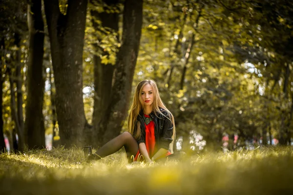 Красивая девушка с длинными волосами в красном платье и кожаной курткой сидит на траве — стоковое фото