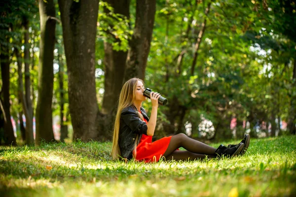 Красивая девушка с длинными волосами в красном платье и кожаной курткой сидит на траве — стоковое фото