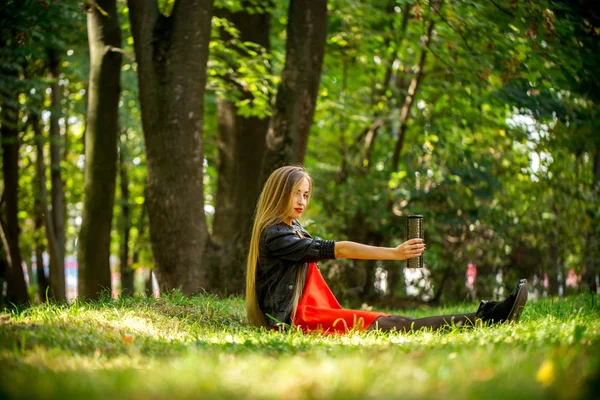 Schönes Mädchen mit langen Haaren in rotem Kleid und Lederjacke auf dem Gras sitzend — Stockfoto