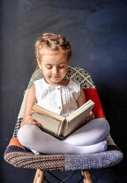 Kleines Mädchen sitzt auf einem Stuhl und liest ein Buch — Stockfoto