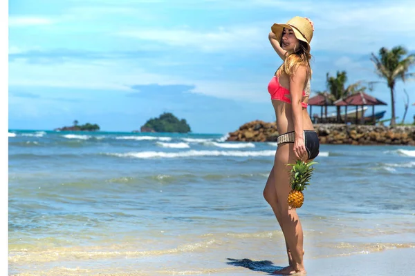 Schönes Mädchen mit Strohhut und wandelnder Ananas an einem exotischen Strand, gutes Essen und Erholung — Stockfoto