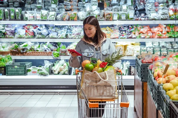Seorang wanita muda membeli bahan makanan di supermarket dengan ponsel di tangannya . — Stok Foto