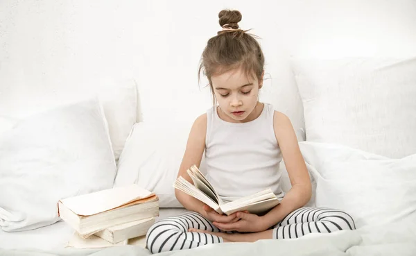 Schattig klein meisje het lezen van een boek op het bed in de slaapkamer. — Stockfoto
