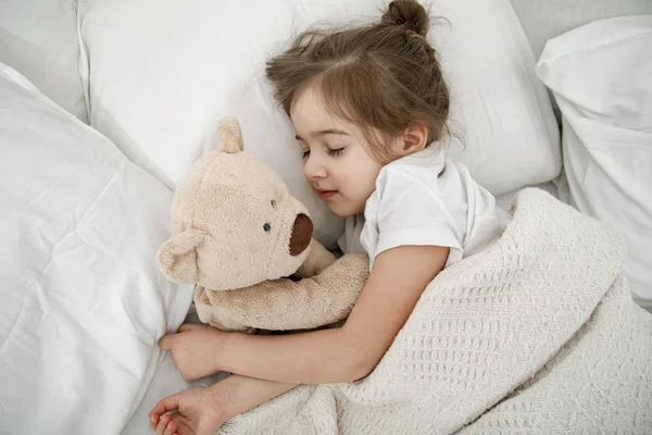 Ein süßes kleines Mädchen schläft in einem Bett mit einem Teddybär-Spielzeug . — Stockfoto