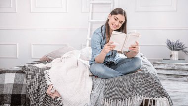 Genç ve güzel bir kadın rahat bir odada yatağın üstünde kitap okuyor. Evde dinleniyorum..