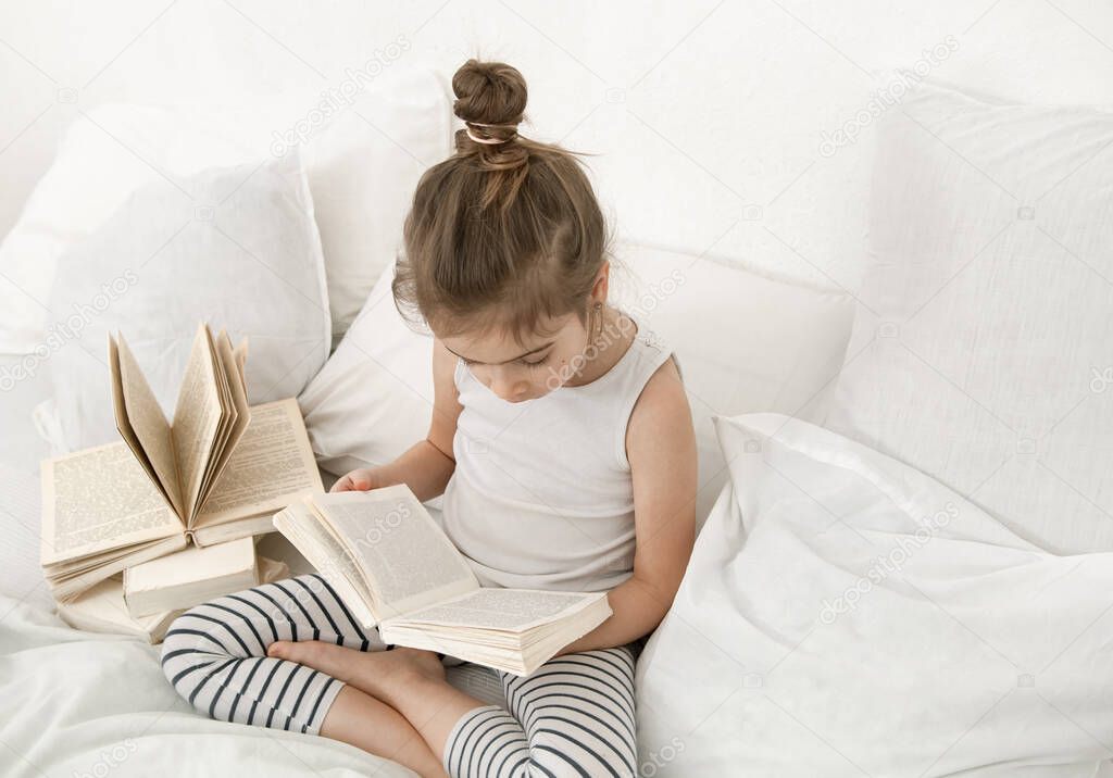Cute little girl reading a book on the bed in the bedroom. The concept of education and family values .