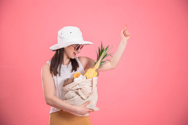 Portrait of a young stylish woman dressed in summer clothes with a hat and sunglasses, holding an ECO-fruit bag, on a pink isolated background.