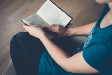 Genç ve güzel bir kadın bir koltuğa oturur ve kitap okur. Açık bir kitap tutan kadın ellerinin yakın çekimi. Konforlu ev