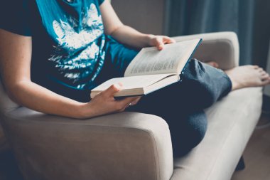 Genç ve güzel bir kadın bir koltuğa oturur ve kitap okur. Açık bir kitap tutan kadın ellerinin yakın çekimi. Konforlu ev