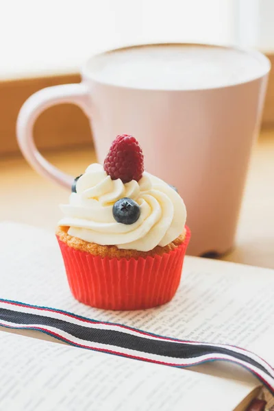 Petit cupcake aux framboises et bleuets sur le livre, tasse de cappuccino chaud — Photo