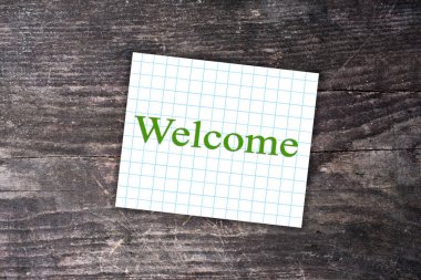 Welcome text on a checkered note. Dark wooden background clipart