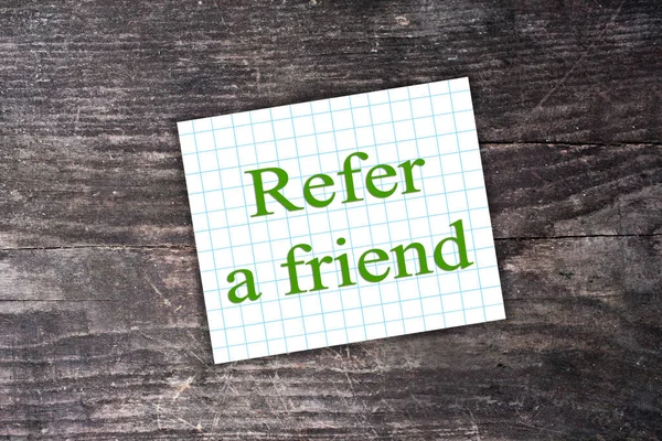 Green Refer a friend text on checkered paper note. Dark Wooden b