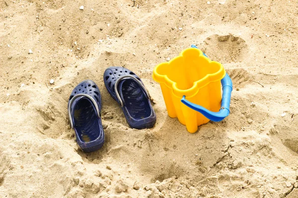 Sapatos de criança e balde de brinquedo amarelo em uma praia, areia. No mar — Fotografia de Stock