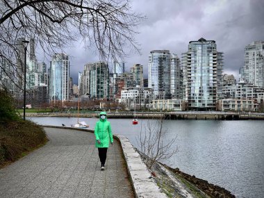 CoVid 19 salgını sırasında deniz kenarında tek başına yürüyen maskeli orta yaşlı bir kadın. Şehir merkezi ve Falls Creek manzarası. Vancouver. İngiliz Kolombiyası. Kanada 