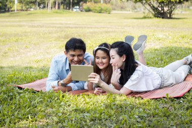 Ebeveynler ve çocuklar parkta paspasın üzerinde tablet çalıyorlar. Annem ve babam tableti kullanarak ders veriyorlar. Kızlar, ebeveynlerin öğrettiği bir tabletten öğreniyorlar..