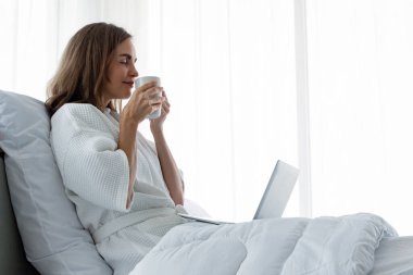 Beyaz pijamalı beyaz bir kadın elinde bir fincan kahve ve içecek tutuyor ve evdeki bir yatak odasındaki laptoptan bilgi alıyor. Kahve içen ve dizüstü bilgisayarda çalışan bir kadın..