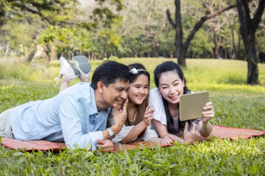Ebeveynler ve çocuklar parkta paspasın üzerinde tablet çalıyorlar. Annem ve babam tableti kullanarak ders veriyorlar. Kızlar, ebeveynlerin öğrettiği bir tabletten öğreniyorlar..