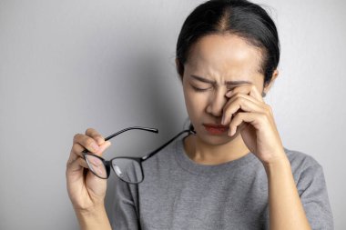 Gri arka planda göz ağrısı olan genç bir kadın. Asyalı kadın göz ağrısı çekiyor. Kadınlar gözlük tutar ve göz ağrısından muzdariptir..