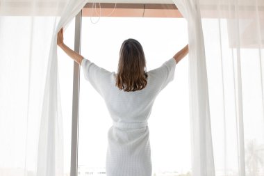 Genç beyaz kadın uyandıktan sonraki sabah pencerenin önünde beyaz perdeleri araladı. Kadın taze bir şekilde uyanır ve pencerelerdeki perdeleri açar..
