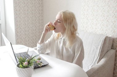 Uzaktan kumandada dizüstü bilgisayar kullanan genç bir kadının yakın çekimi. Beyaz gömlekli bir kız. İş yerindeki sıkıcı kadın elma yiyor. Seçici odak.