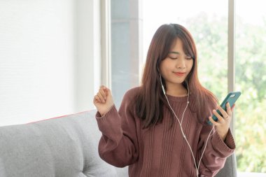 Güzel Asyalı kadın oturma odasındaki gri koltukta akıllı telefon kullanıyor.