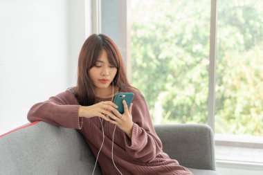 Güzel Asyalı kadın oturma odasındaki gri koltukta akıllı telefon kullanıyor.