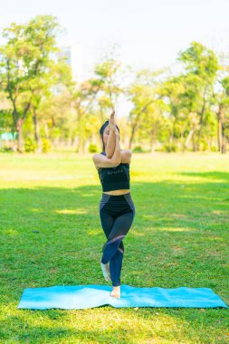 Asyalı kadın yoga yapıyor ve yazın yeşil parkta spor kıyafetleriyle rahatlıyor.