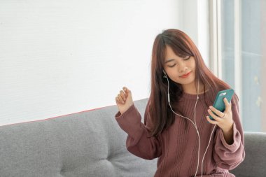 Güzel Asyalı kadın oturma odasındaki gri koltukta akıllı telefon kullanıyor.