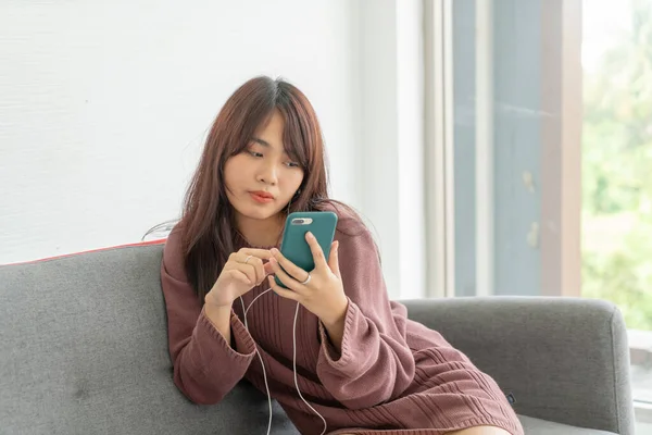 在客厅的灰色沙发上使用智能手机的漂亮亚洲女人 — 图库照片
