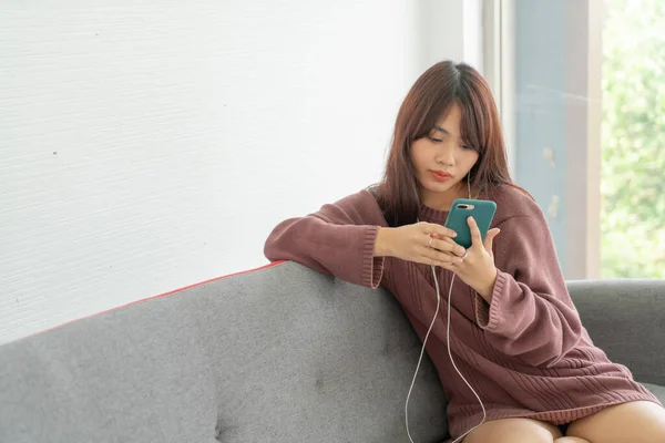 在客厅的灰色沙发上使用智能手机的漂亮亚洲女人 — 图库照片