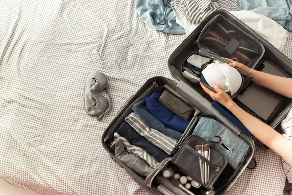 Female hands packing traveler case on bed, closeup. Beach accessories in opened suitcase. Travelling wheeled bag, different clothes.