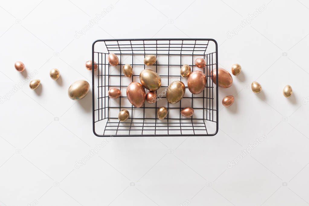 Top view a metal basket stands on white isolated background with golden copper and black and white minimalistly painted eggs. Easter and stylish decor concept. Advertising space